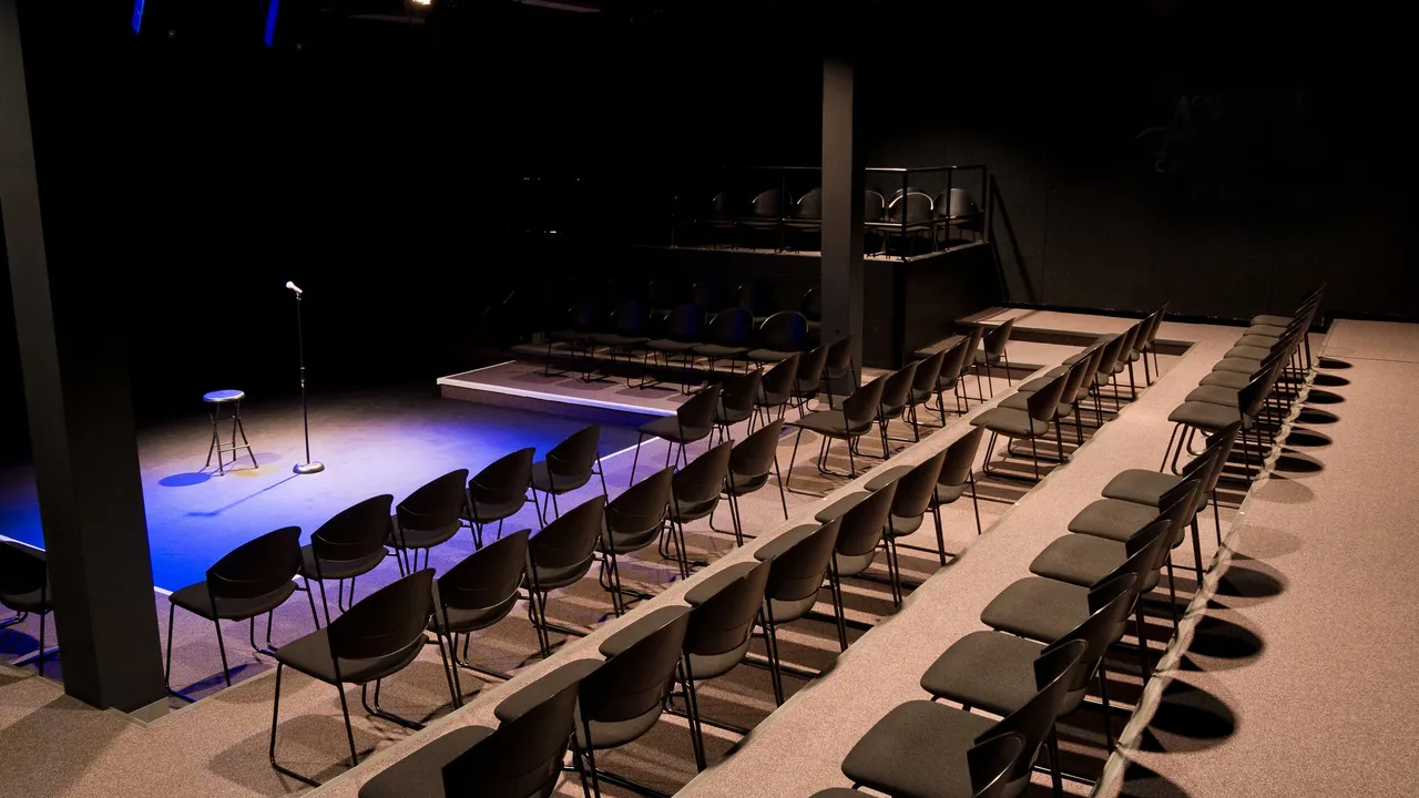 An empty black-box theater with rows of chairs facing a small lit stage