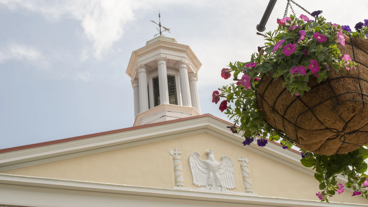 A white cupola and an eagle relief atop a historic building beside a hanging flower basket
