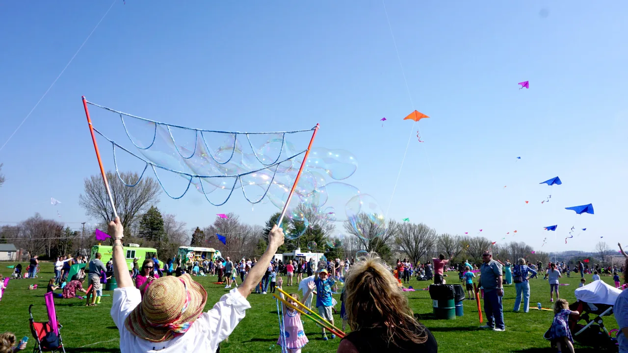 Children and families fly kites and wave giant bubble wands in a grassy park