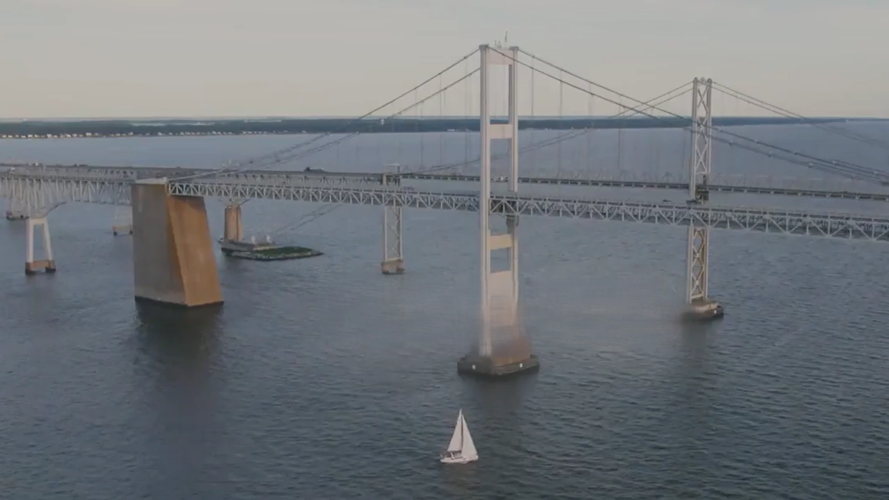 A long suspension bridge spans the bay with a small sailboat on the water below.