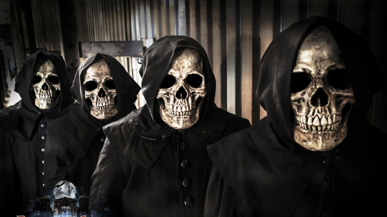 Four figures in black hoods and skull masks stand together in a dark room.