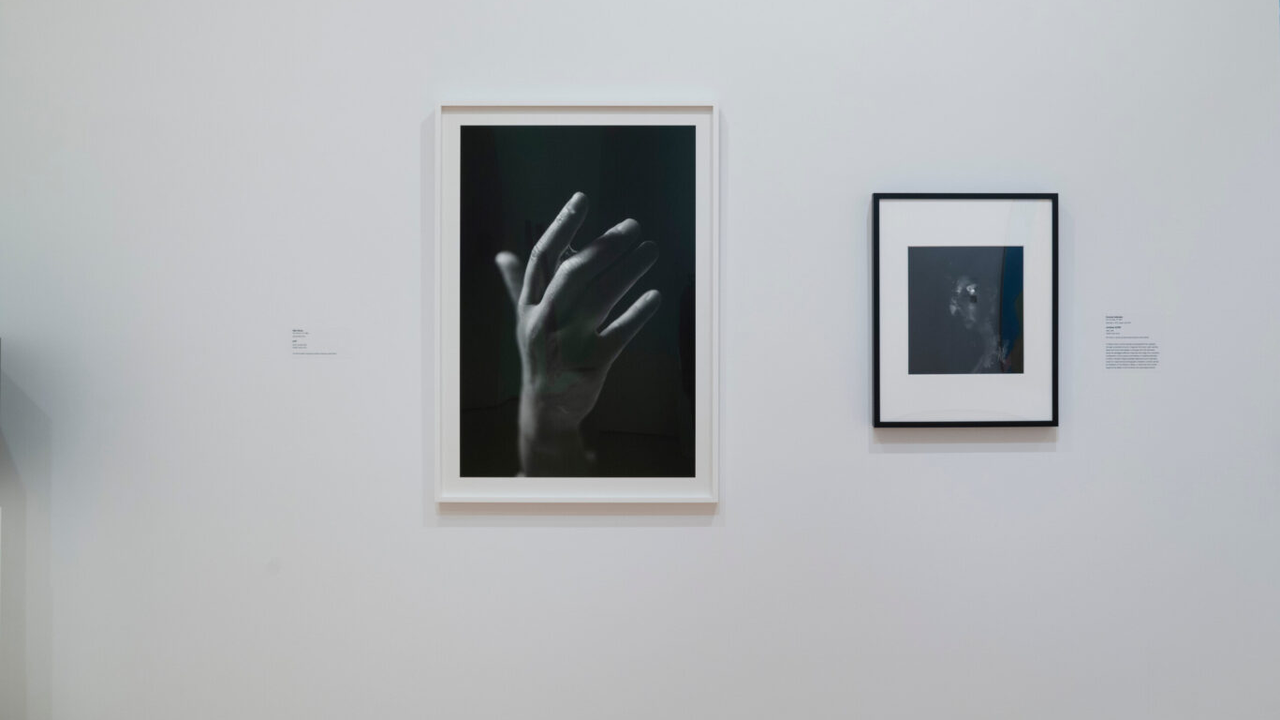 Two framed black-and-white photographs, one of a raised hand, hang on a white gallery wall.