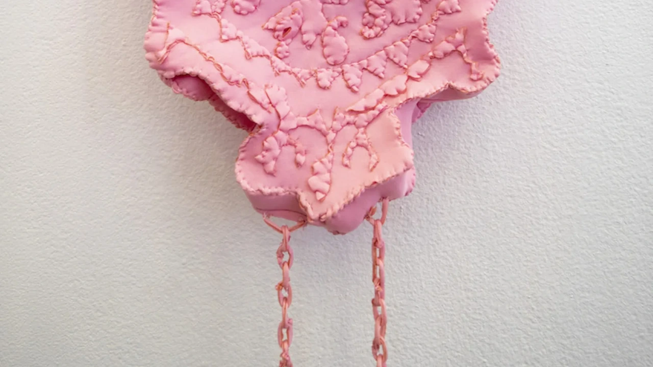 A pink lace-textured fabric sculpture hangs from a white wall by pink threads.