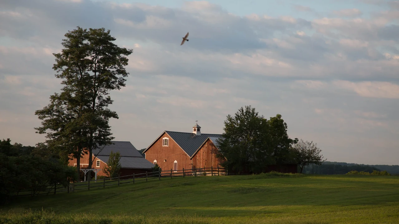 A red brick farmhouse and barn on a green hillside under a soft evening sky.
