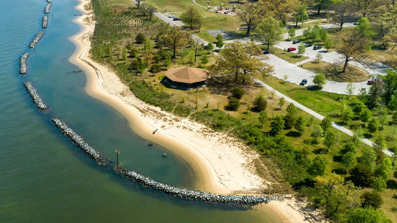 Aerial view of a sandy point with stone breakwaters, trees and walking paths.