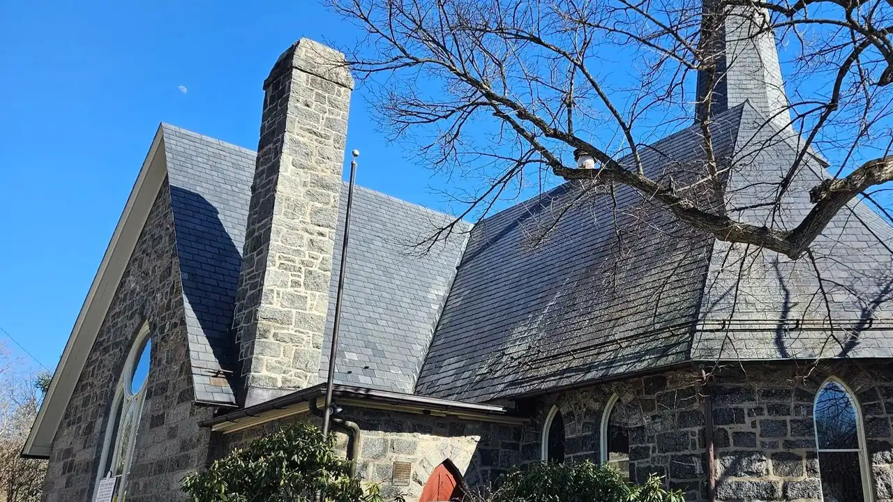 A gray stone church building with a tall steeple and a bare tree against a blue sky.
