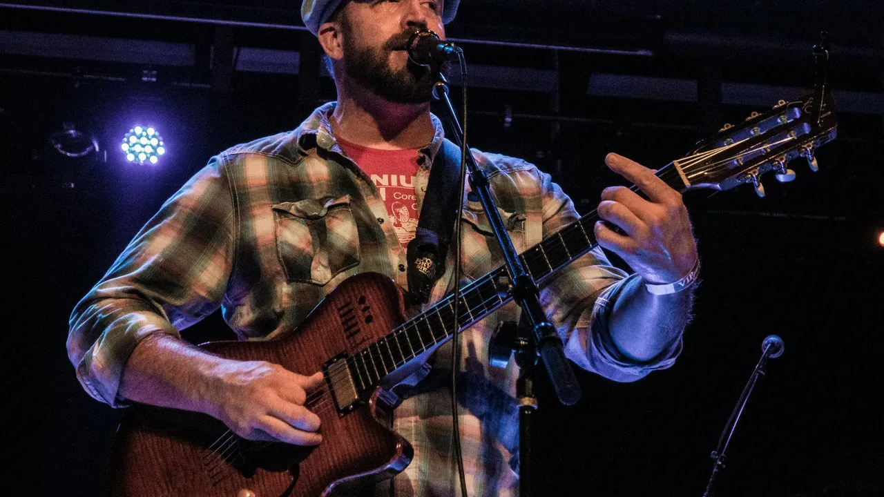 A bearded guitarist in a plaid shirt sings into a microphone on a dark stage.
