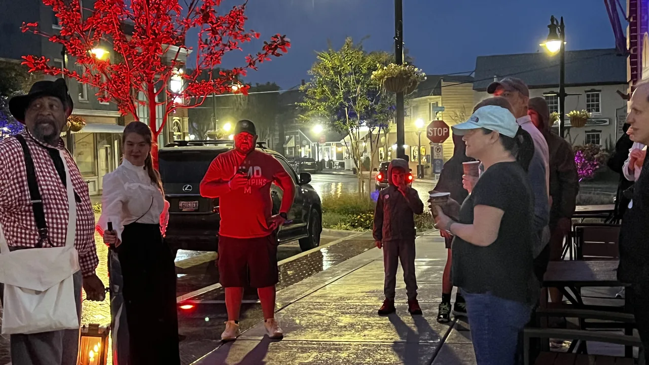 A guide in a plaid shirt and hat speaks to a small group on a wet street at night.