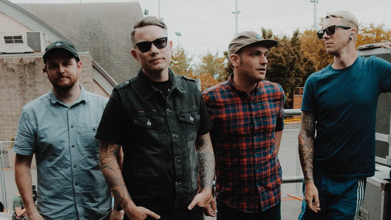 Four men from a band stand together outdoors, two in sunglasses.