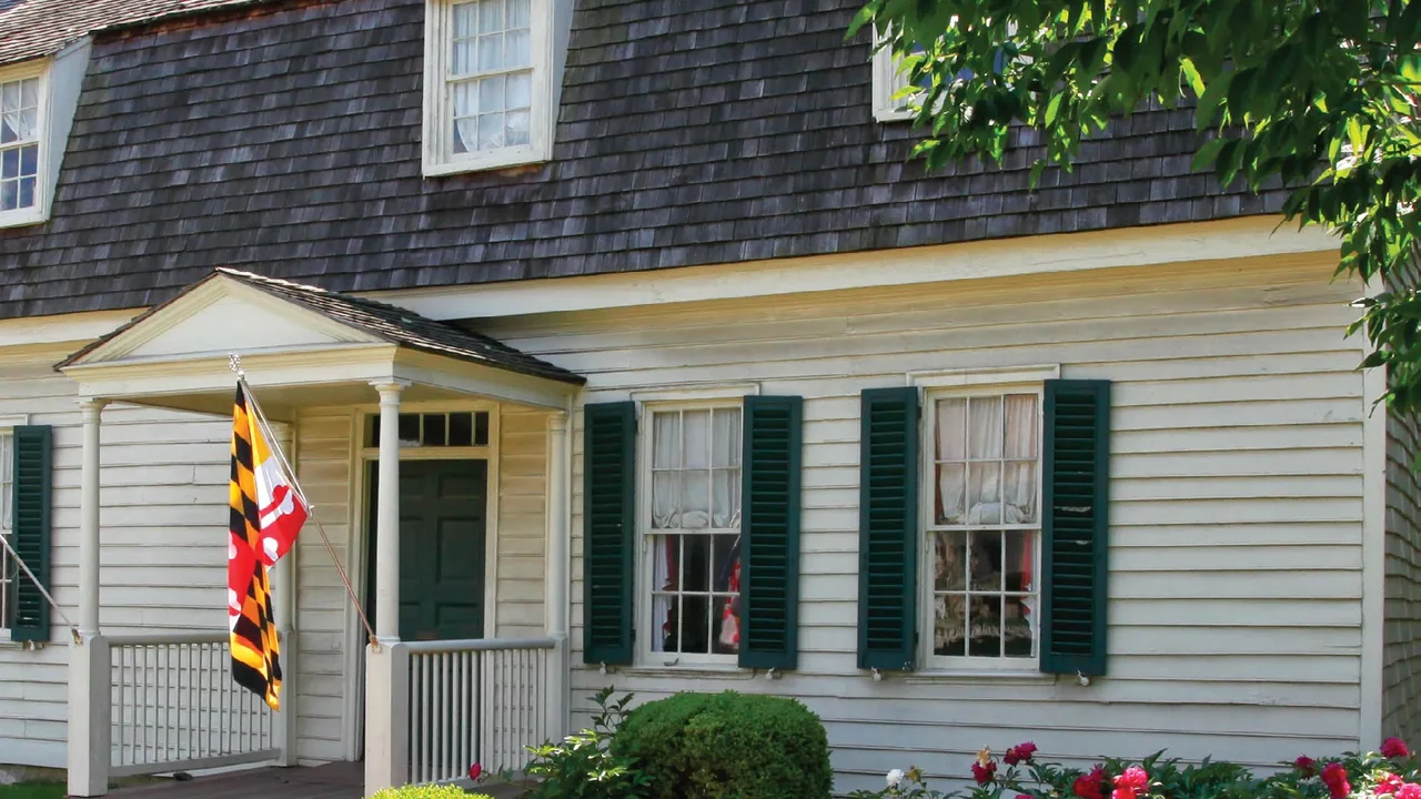 A white clapboard colonial house with green shutters, a small porch and a Maryland flag.