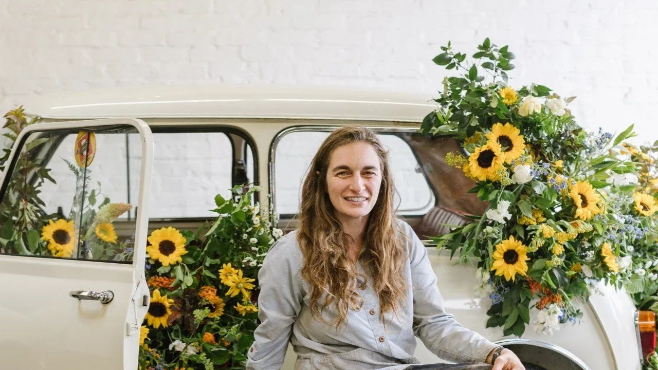 A smiling woman sits in a vintage cream car decorated with sunflowers.