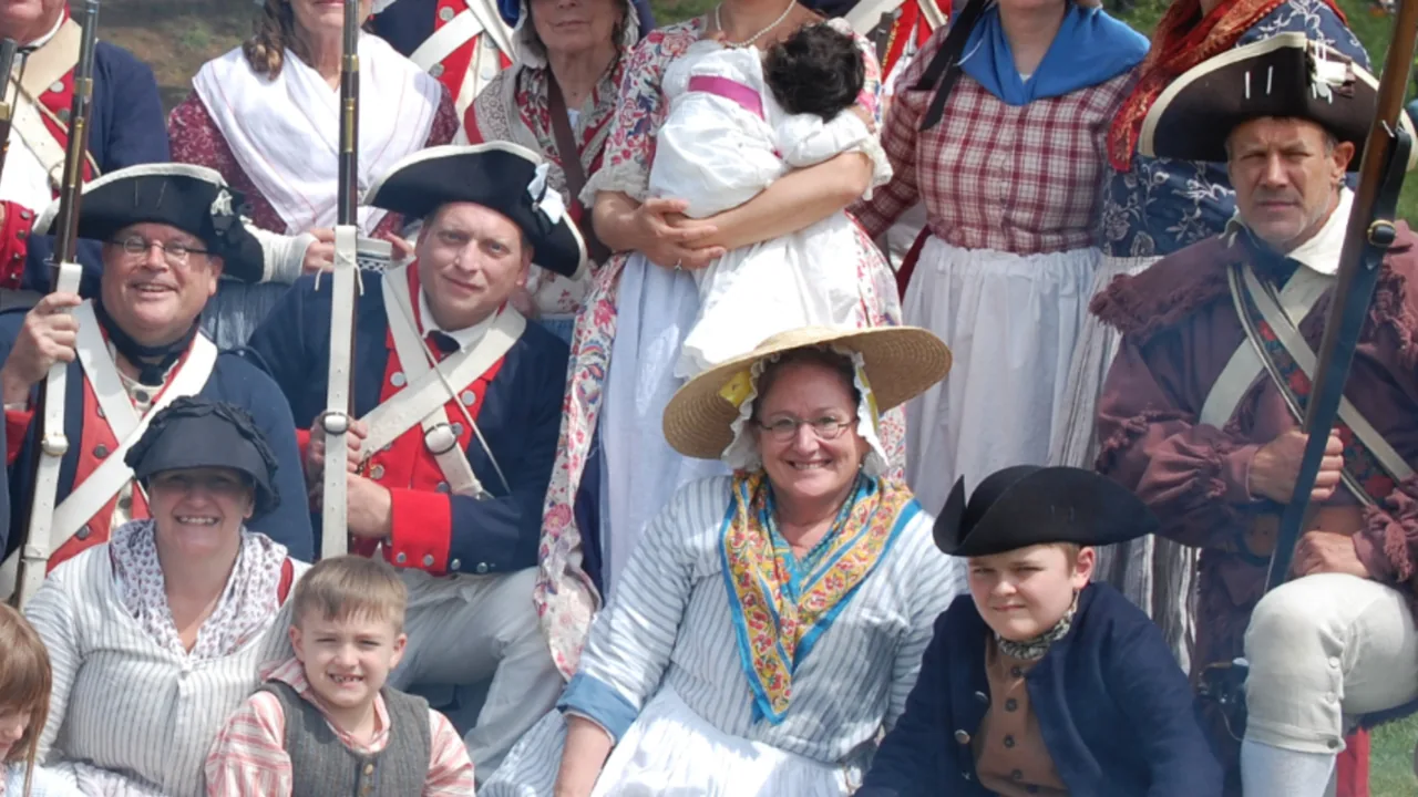Reenactors in colonial uniforms and dresses gather with children for a group photo.
