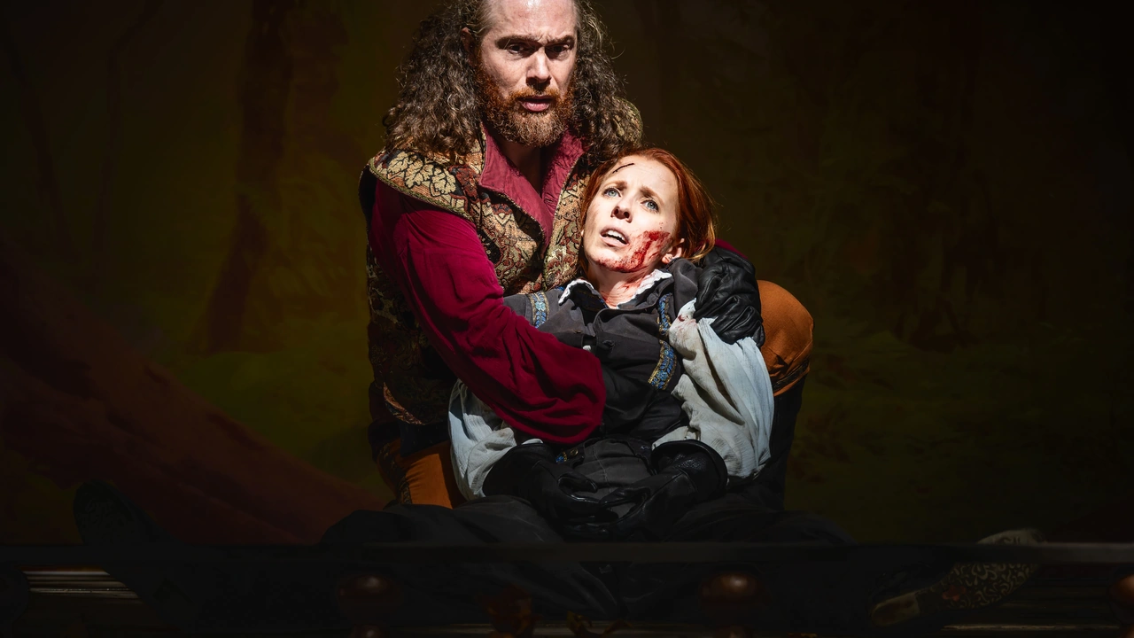 A bearded actor in a red costume cradles a red-haired woman on a dark stage.