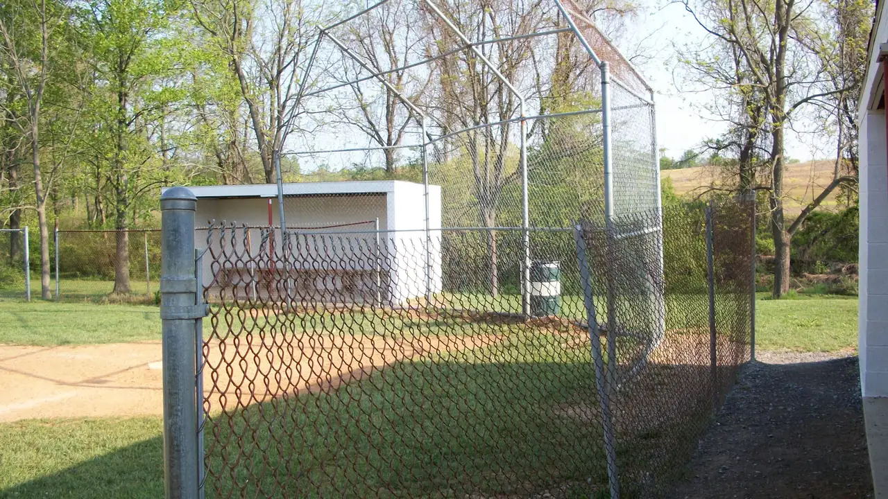 A fenced ball field with a backstop and a small white shed among trees