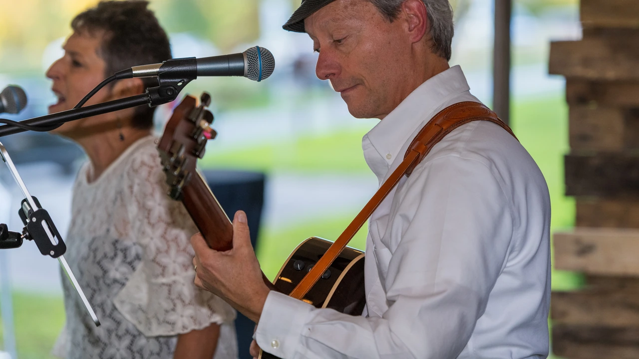 A woman sings and a man plays acoustic guitar at microphones in an open-air pavilion.
