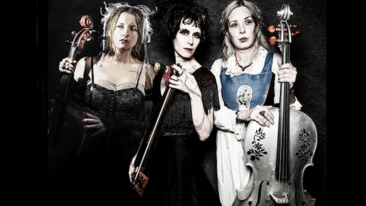 Three musicians in dark dresses hold cellos and pose against a black backdrop