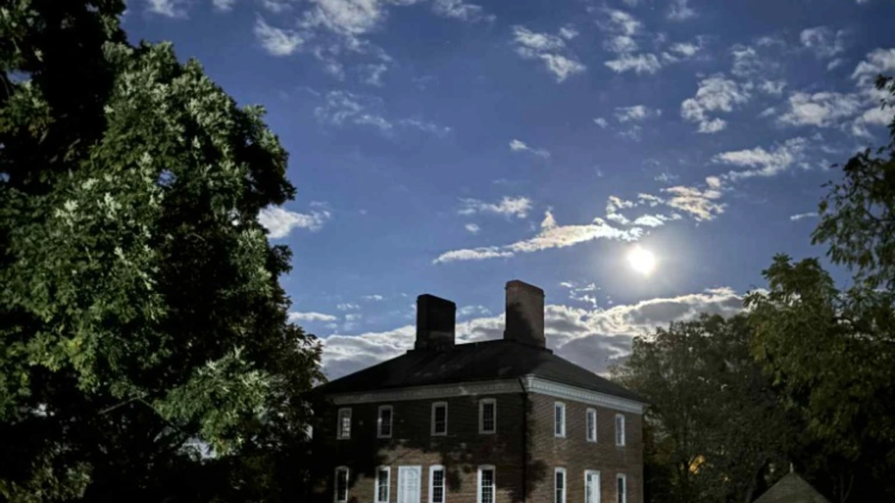 A brick colonial house beneath trees with a bright moon in a dusky sky.