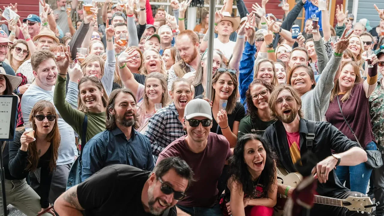 A large cheering crowd poses for the camera behind a band at an outdoor festival.