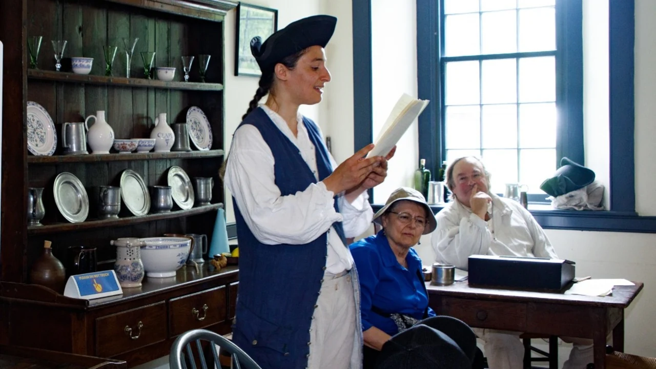 An interpreter in colonial dress reads from a paper to seated visitors in a historic room.