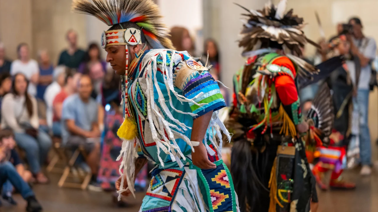 Dancers in feathered regalia and bright ribbon outfits perform before a seated audience.