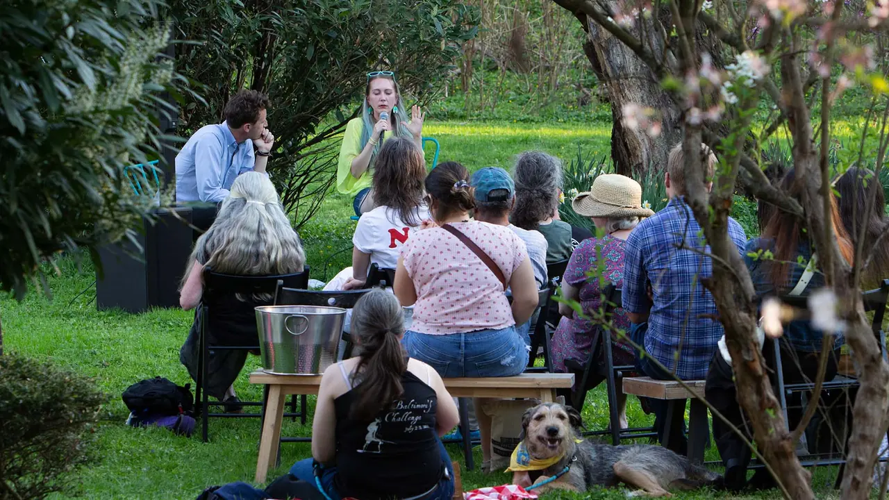People sit on chairs and benches on a sunny lawn under trees, a dog at their feet