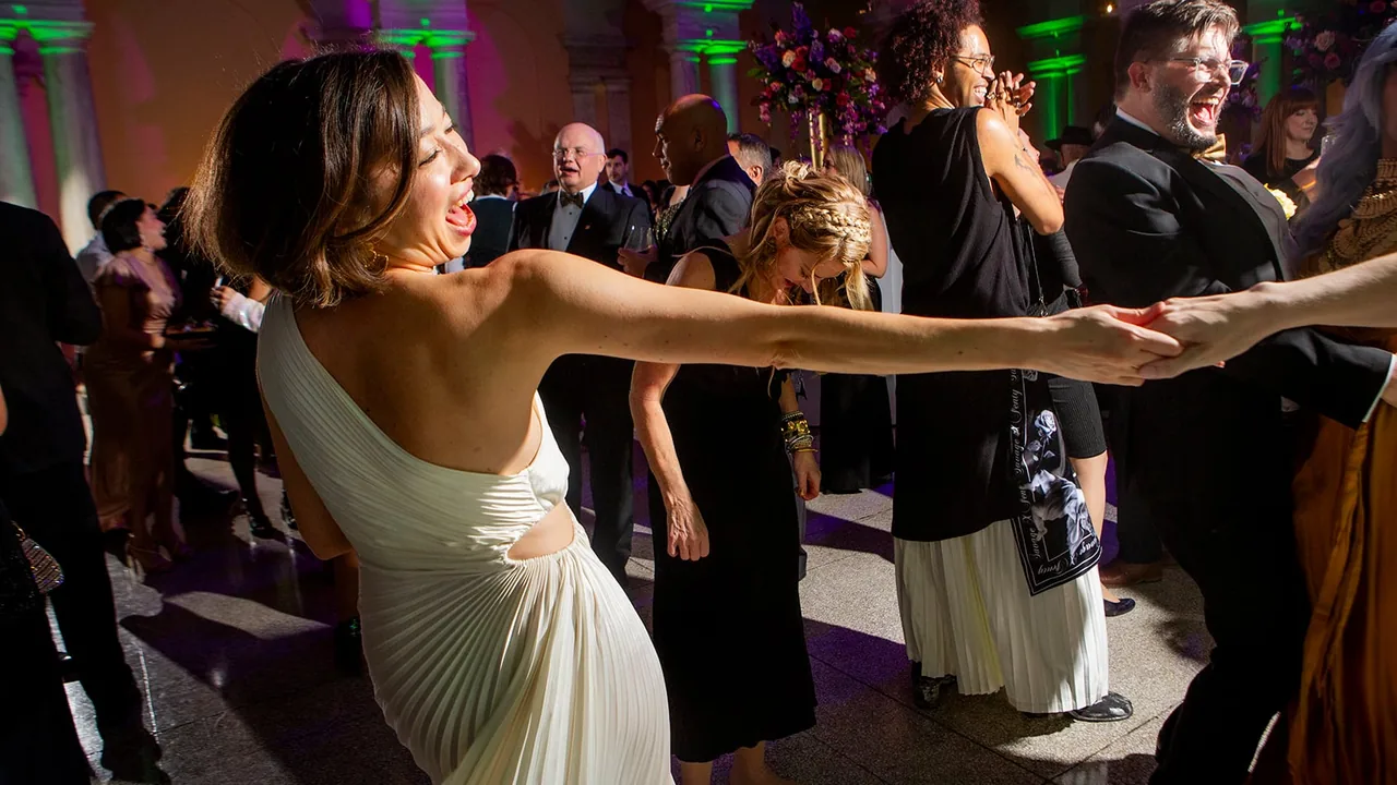 A woman in a white gown laughs while dancing with a partner among guests at a party.