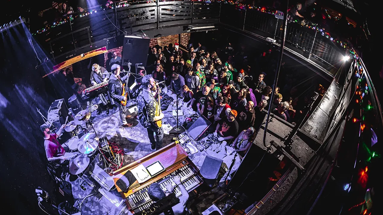 A band plays on a stage lit in blue as a crowd fills the floor of a small club, seen from above
