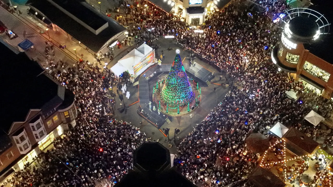 An overhead night view of a packed plaza surrounding a lit Christmas tree, ringed by shops.