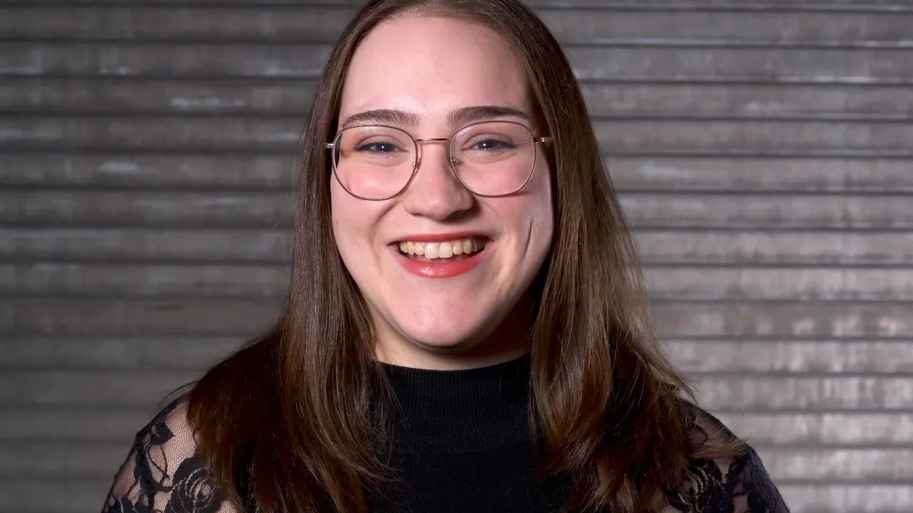 A smiling woman with long brown hair and glasses in front of a gray wooden wall.