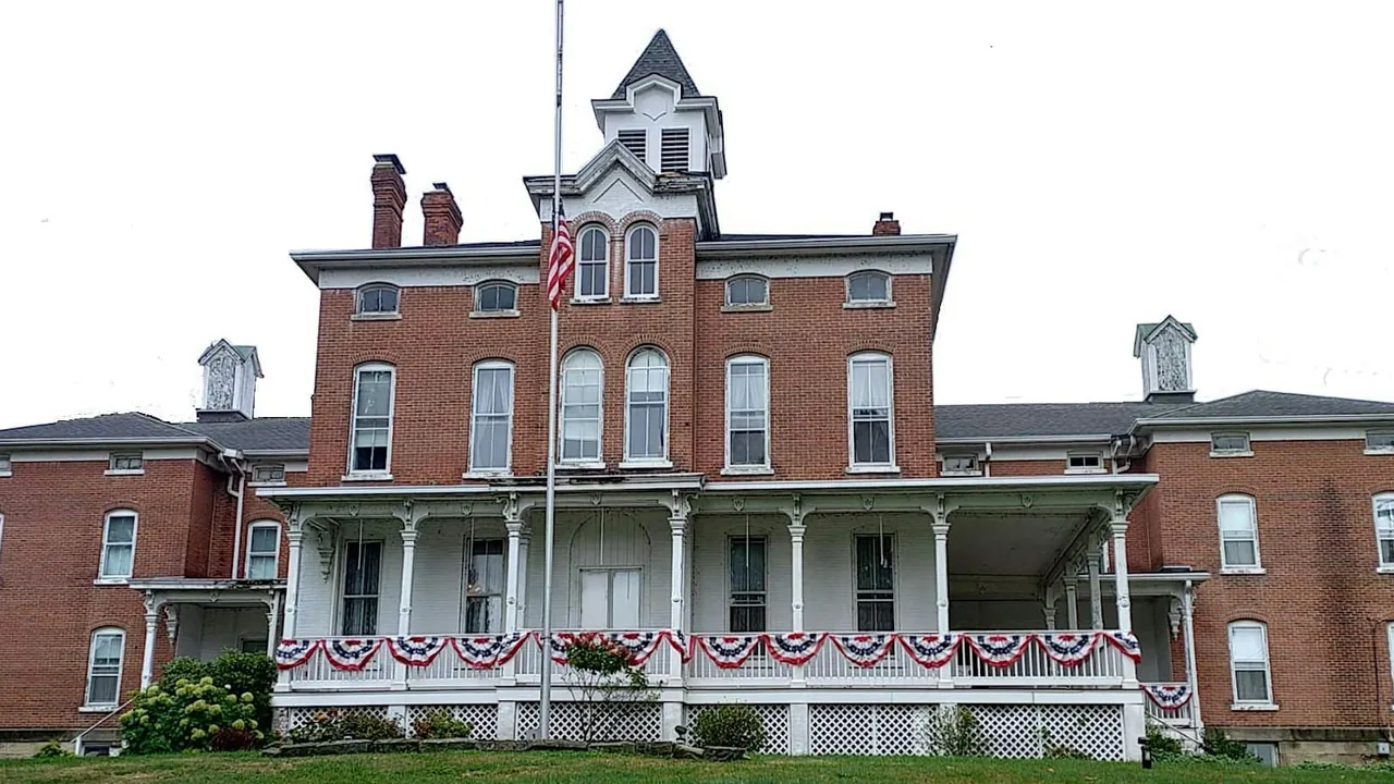 A three-story brick mansion with a cupola, white porch and red, white and blue bunting under an overcast sky.