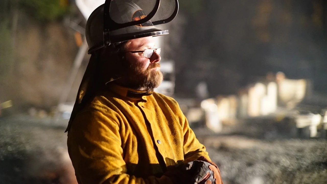 A bearded man in a welding helmet and protective jacket stands near molten-metal work.