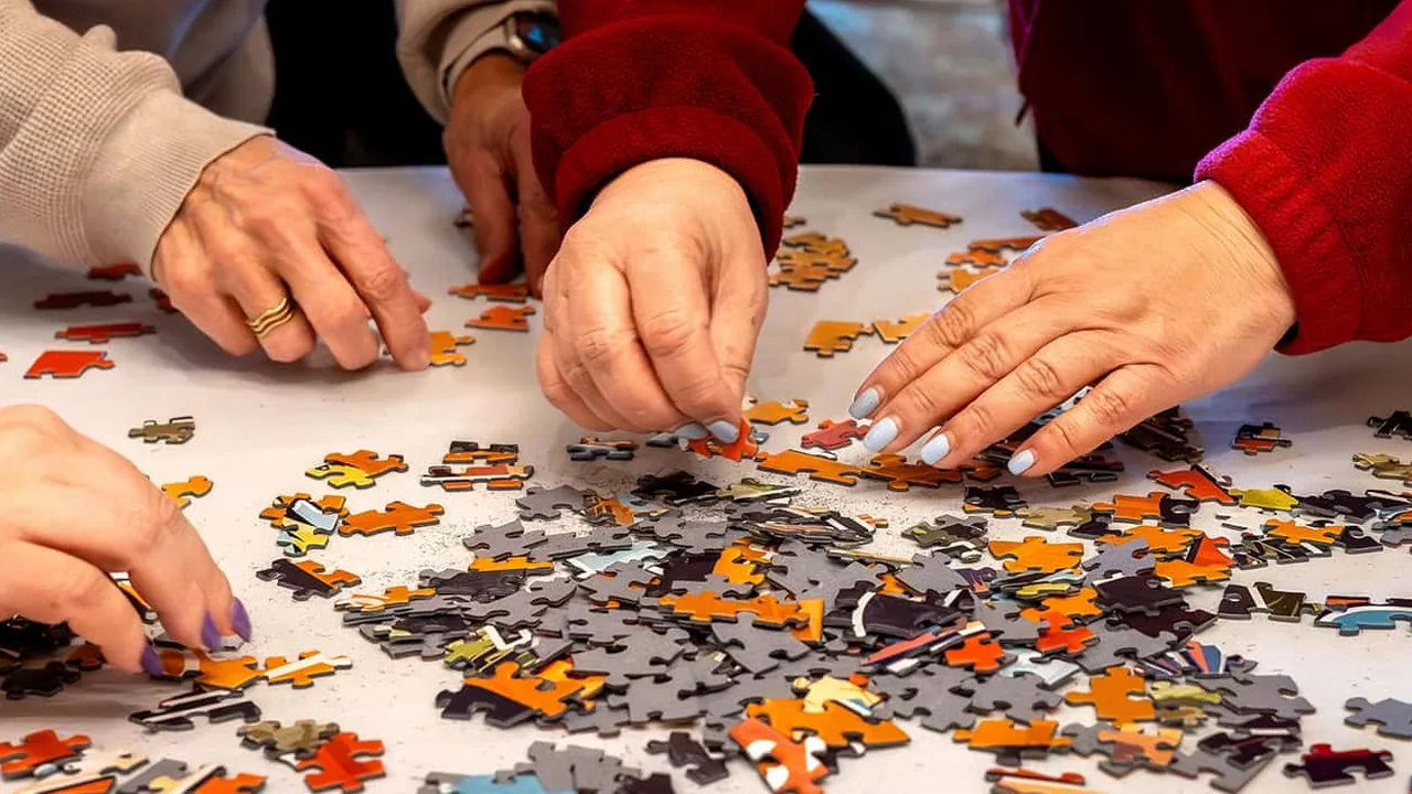 Several hands assemble a jigsaw puzzle with bright orange and gray pieces on a table.