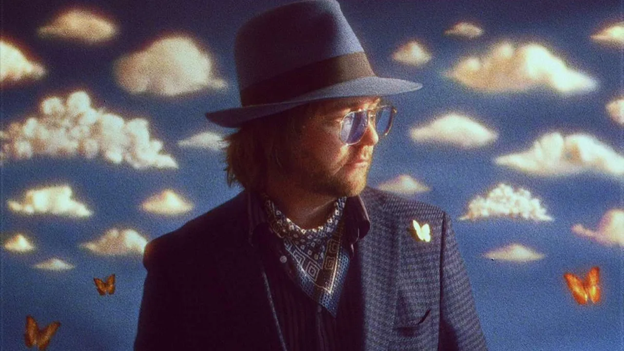 A man in a fedora and glasses stands before a painted backdrop of clouds and butterflies.