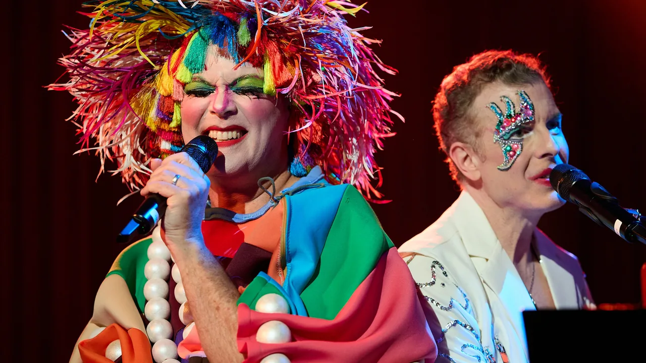 Two performers in colorful wigs and face paint sing into microphones on stage.