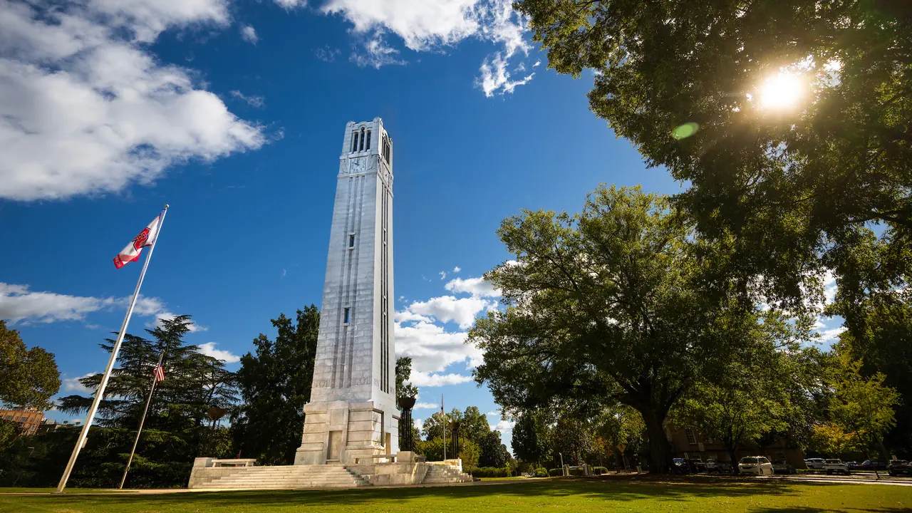 A tall stone bell tower and a flagpole on a green lawn under a blue sky.