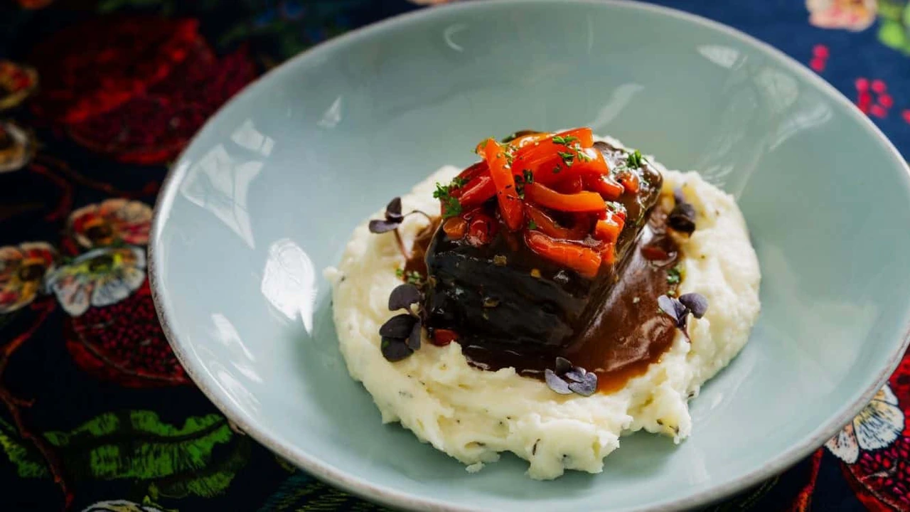 A plated dish of braised meat with roasted peppers over mashed potatoes in a pale blue bowl.