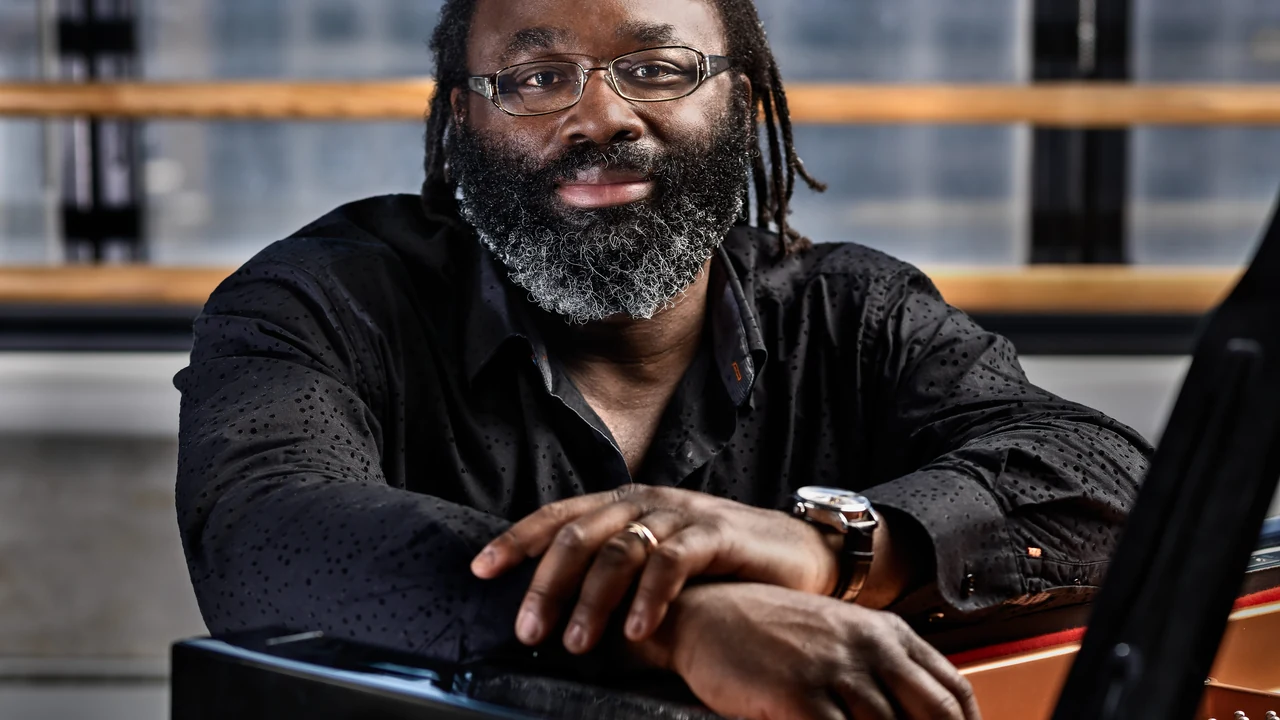 A pianist with glasses and a beard rests his arms on a piano and smiles.