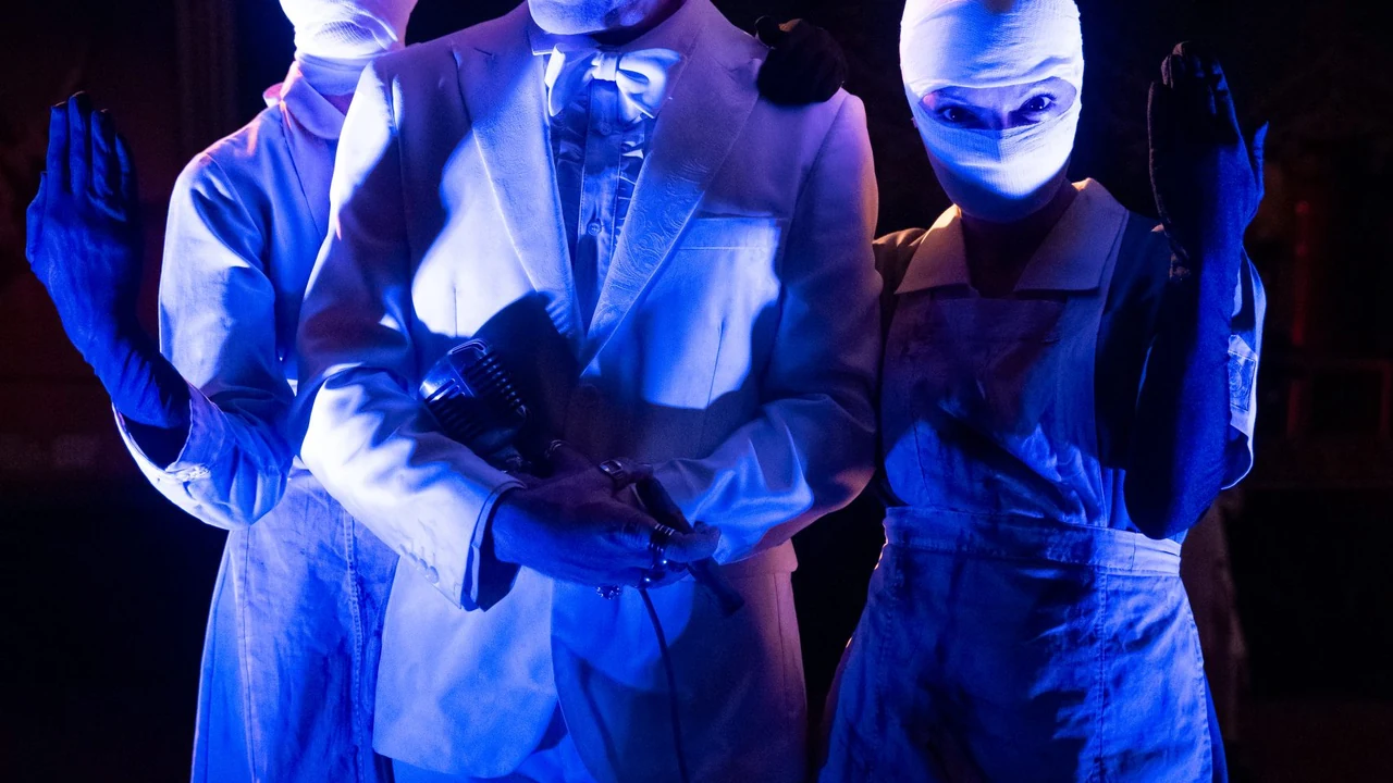 Three performers in white masks and pale costumes lit in blue on a dark stage.