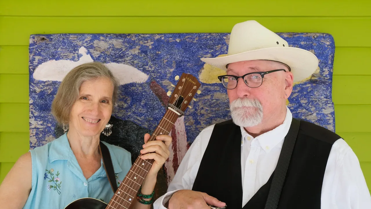 A duo with guitar and cowboy hat pose before a painted backdrop