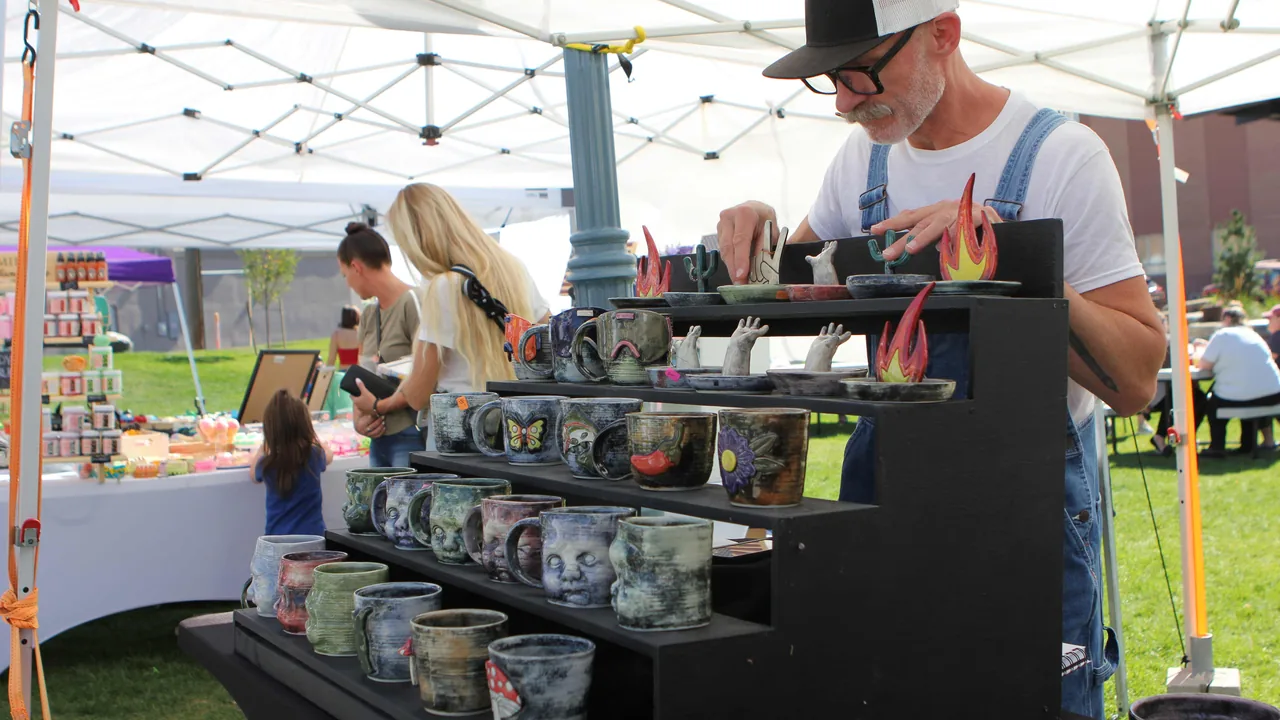 A potter in a hat arranges colorful mugs on a stand at an outdoor market tent.