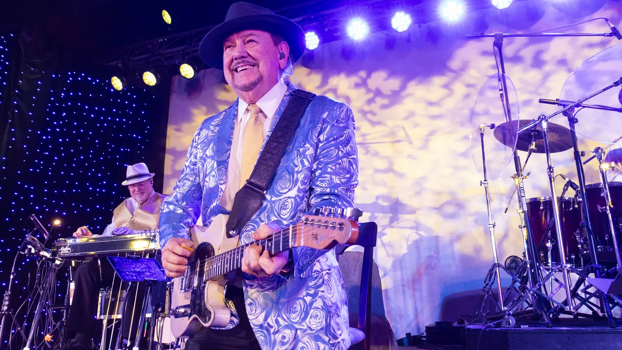 A guitarist in a cowboy hat and patterned blue suit performs on a stage with a band.