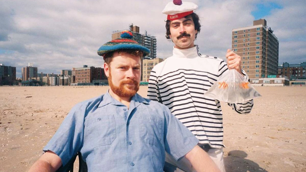 Two men in quirky hats stand on a city beach, one holding a bag with a goldfish.