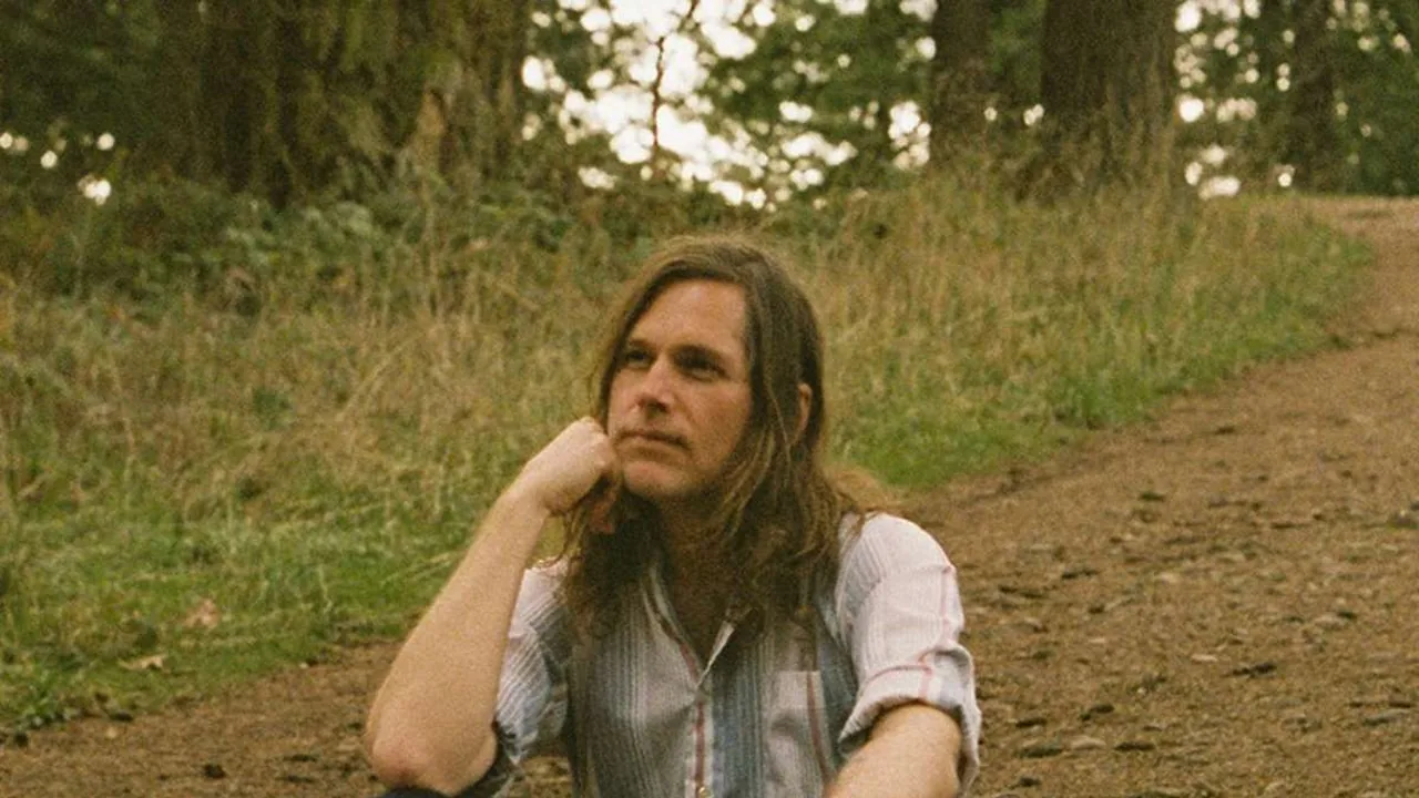 A long-haired man sits beside a dirt path at the edge of a forest.