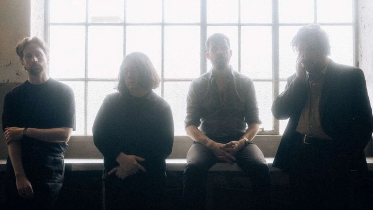 Four band members sit in silhouette in front of a bright window.
