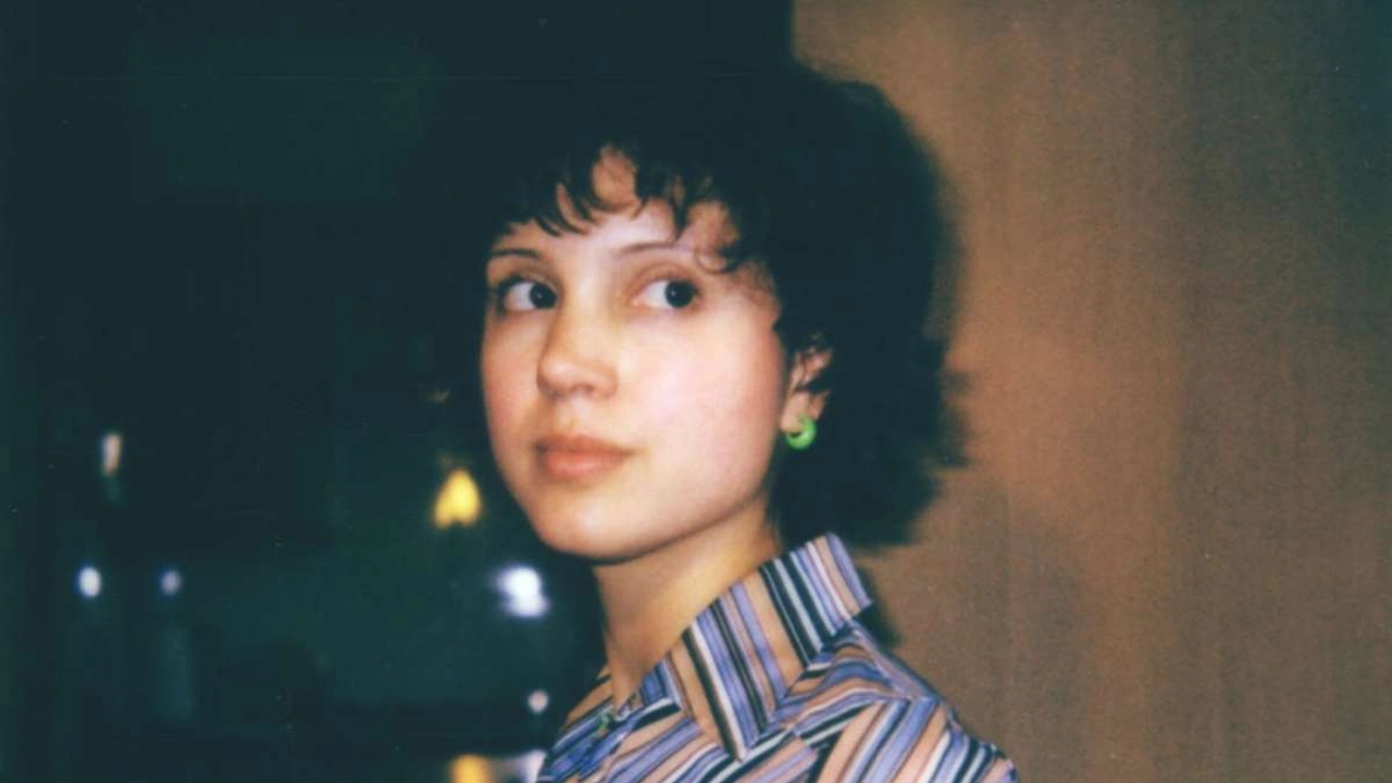 A young woman with dark curly hair and a striped collar looks toward the camera.
