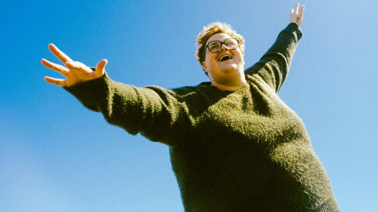 A smiling person in a green sweater throws their arms wide against a blue sky.