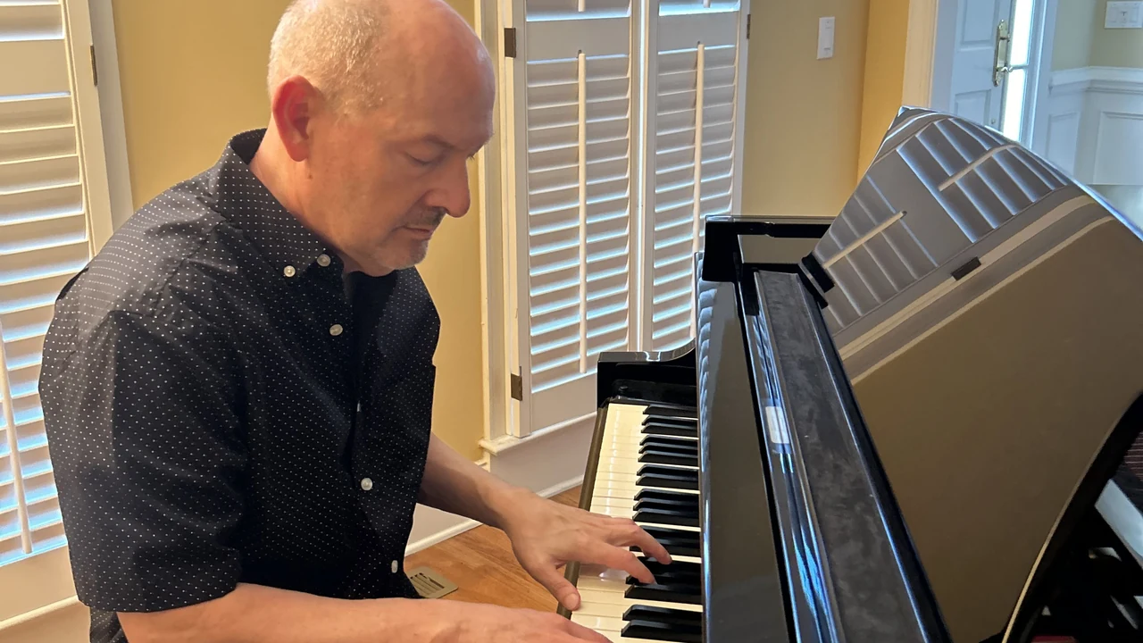 A man in a dark shirt plays a grand piano in a sunlit room.