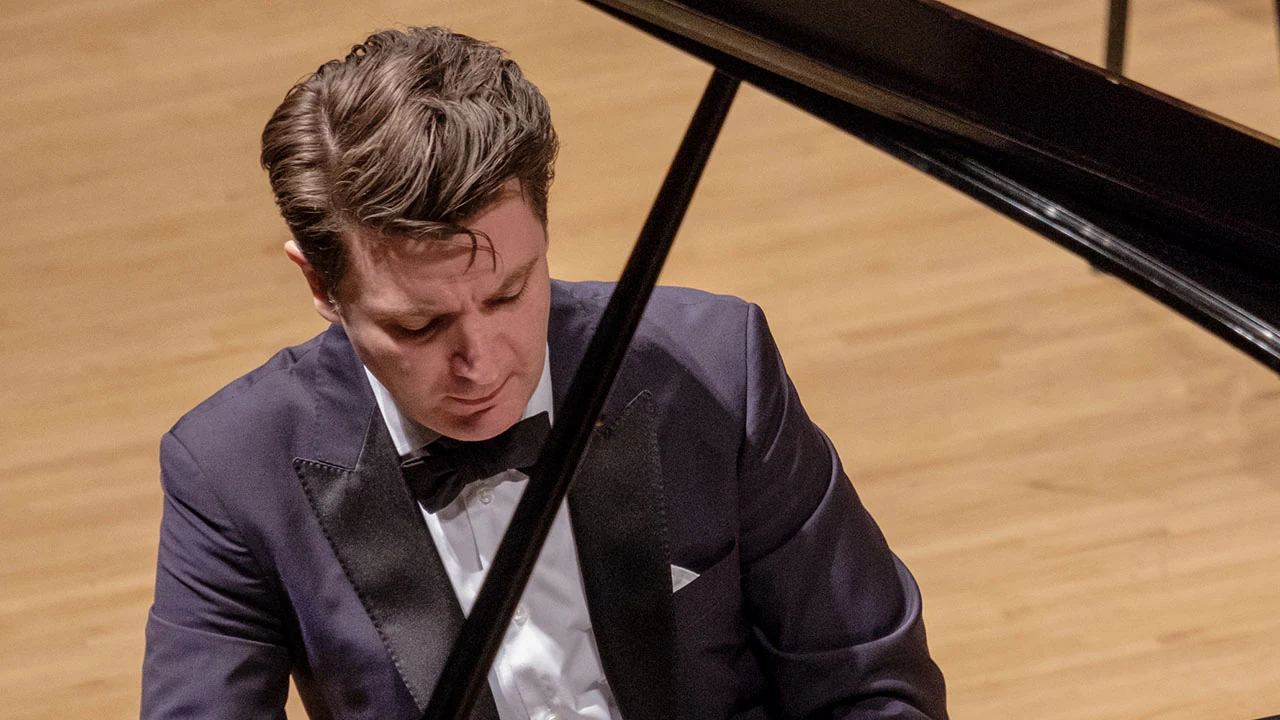 A pianist in a tuxedo plays a grand piano on a wooden stage.