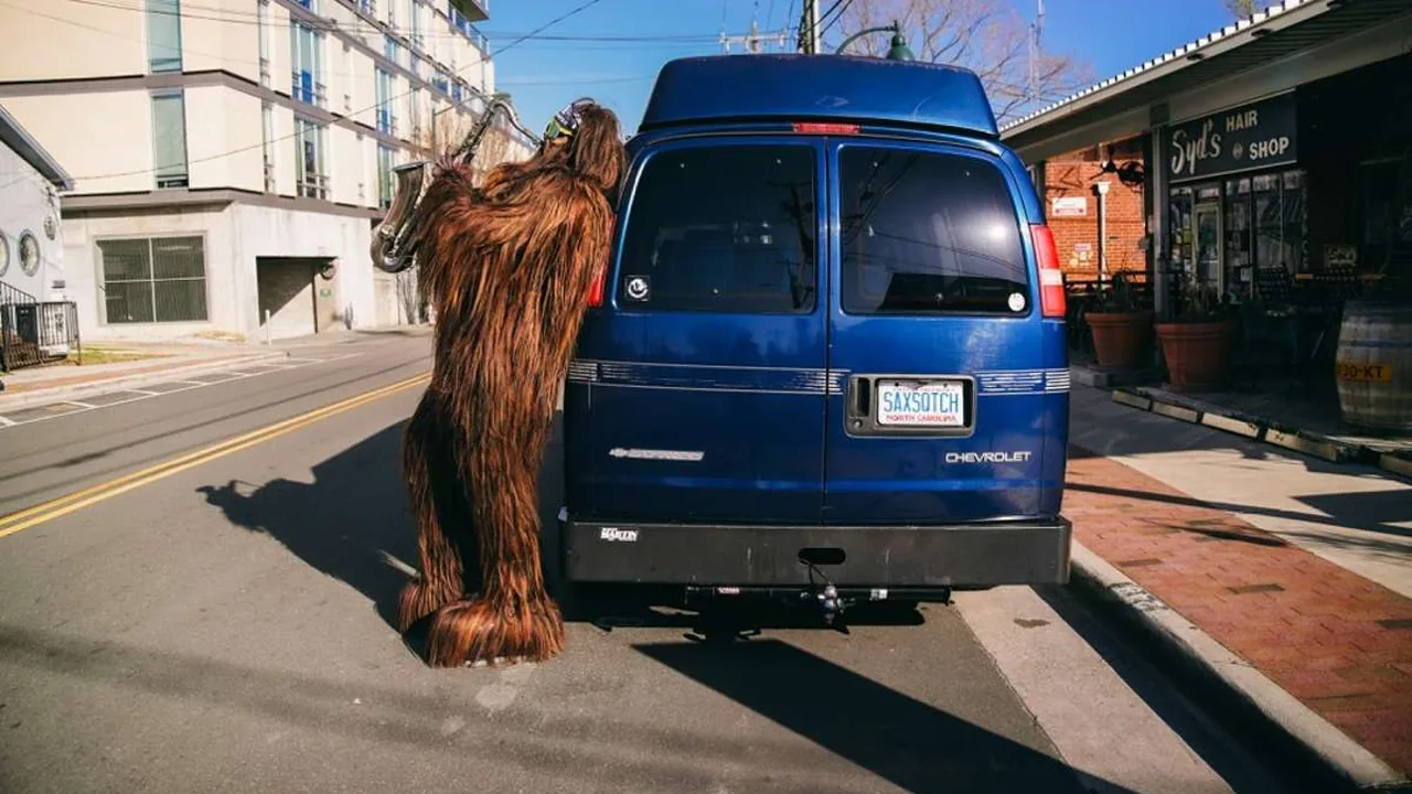 A person in a shaggy brown creature costume stands beside a blue van on a street.