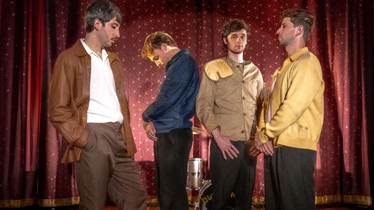 Four men in jackets perform a scene on a stage with red curtains.